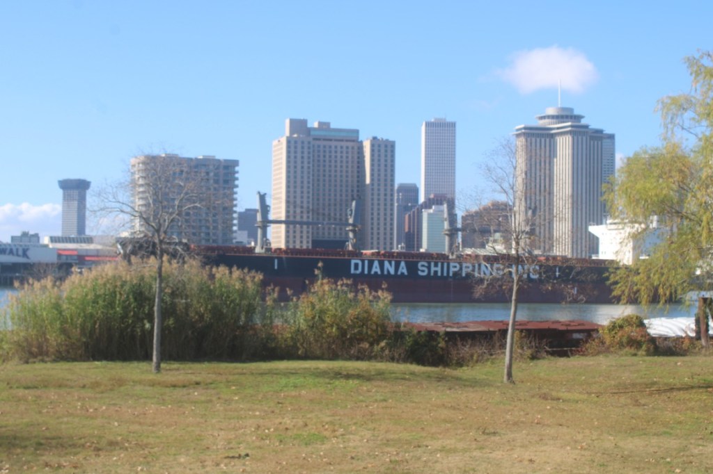 A photograph of a large merchant ship transiting the Mississippi with the New Orleans skyline in the background.
