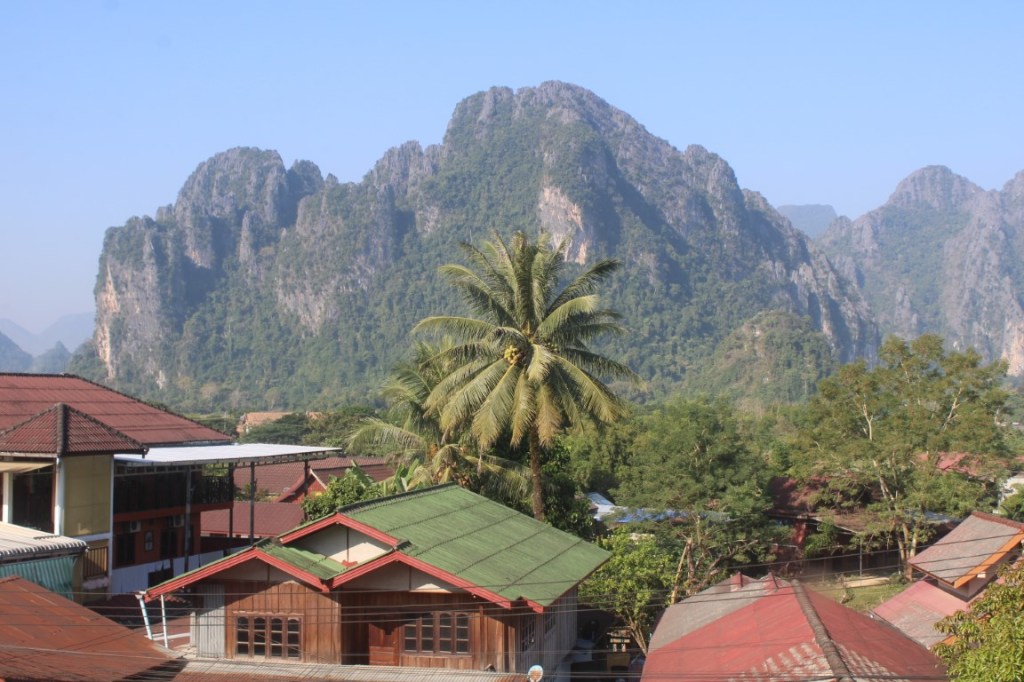 Photograph with Vang Vieng Village and a palm tree in the foreground, and karst mountains in the background. 