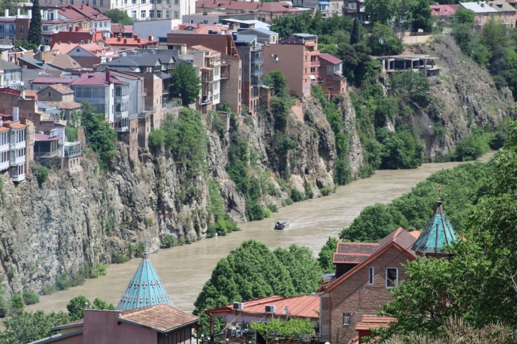 Photograph showing the Kura River flowing through Tbilisi, Georgia with bank on one side and cliff on the other. 